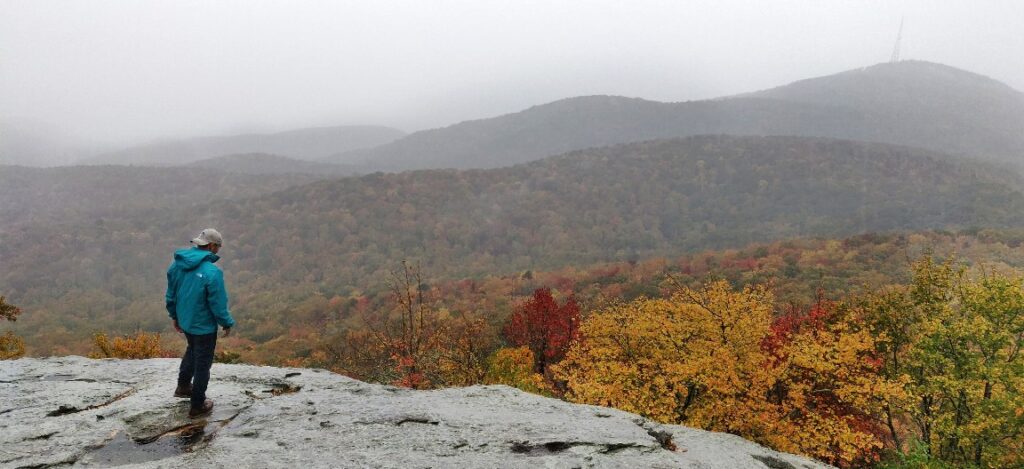 man in teal rainjacket standing on wet rocky ledge. Background: foggy valley of trees changing into fall colors
