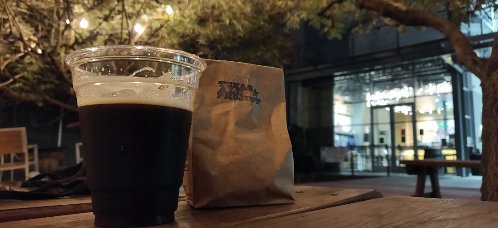 Foreground: plastic cup filled with dark beer & brown paper bag of peanuts on picnic table. Background: floor-to-ceiling glass windows looking into brewery.