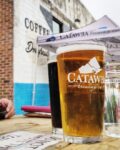 Pint glass full of beer on outdoor picnic table. Background: exterior of Catawba's South Slope taproom