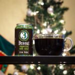 A clear glass coffee mug filled with 3 Nation's Imperial Milk Stout. On a table next to an empty can from 3 Nations Brewing. Background: out-of-focus Christmas tree