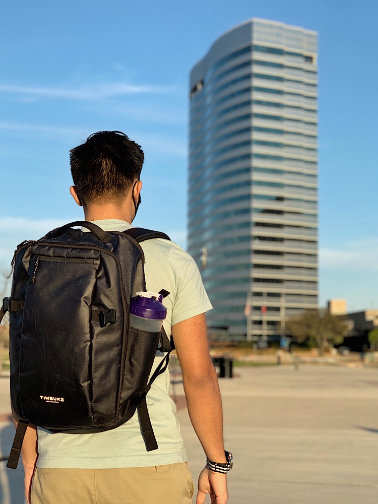 Man wearing jet black Timbuk2 Blink Pack backpack outside. Tall business tower in background.