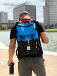 Man wearing royal/black Topo Designs Rover Pack while walking along an urban lake